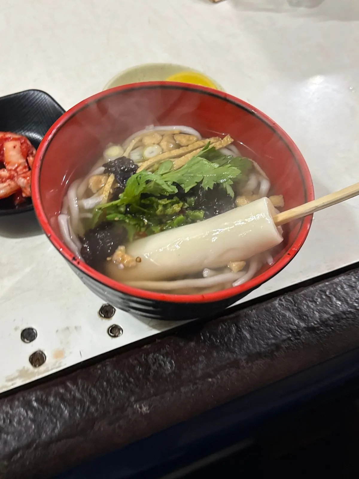 붉은 그릇에 담긴 우동과 그 위에 올린 꼬치 떡, 오뎅