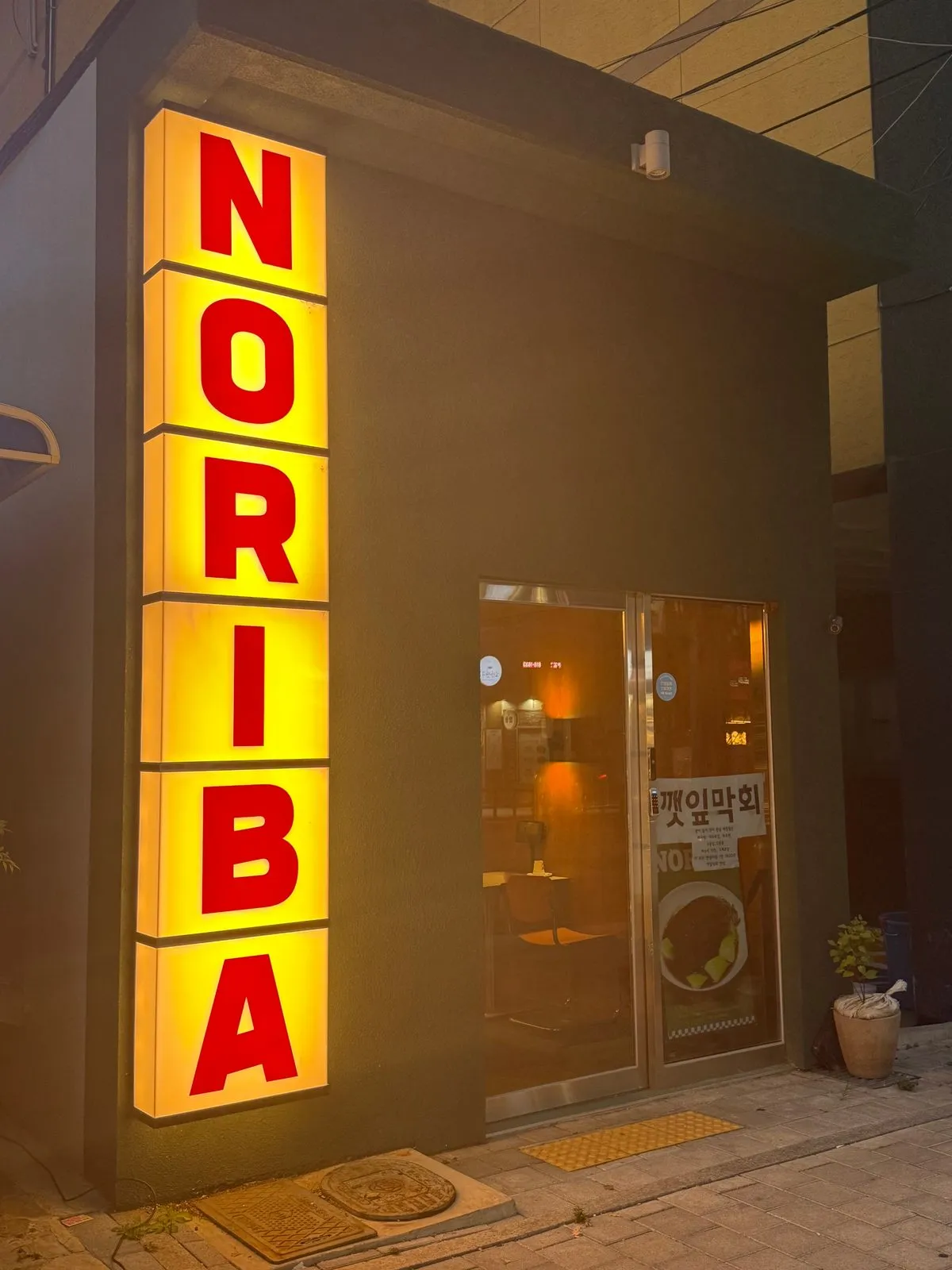 어두운 건물 벽에 세로로 달린 노란 NORIBA 간판과 입구