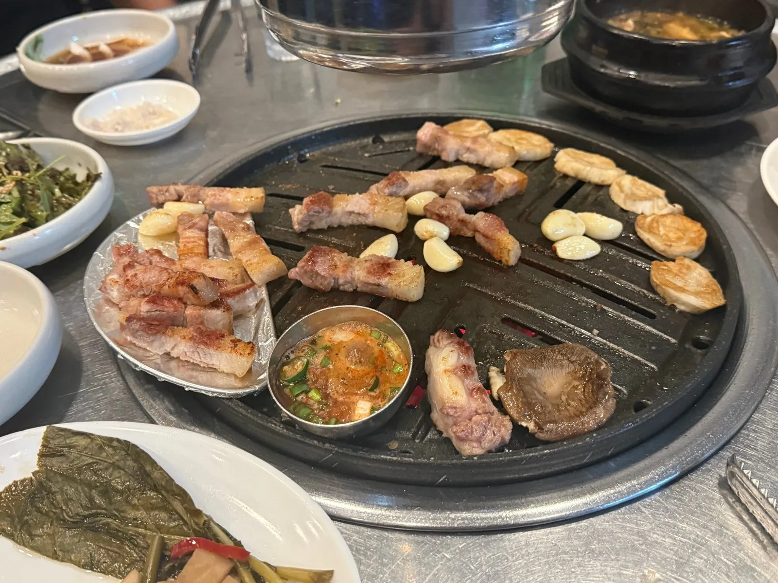 둥근 불판 위에서 익고 있는 삼겹살과 마늘, 그 옆의 반찬