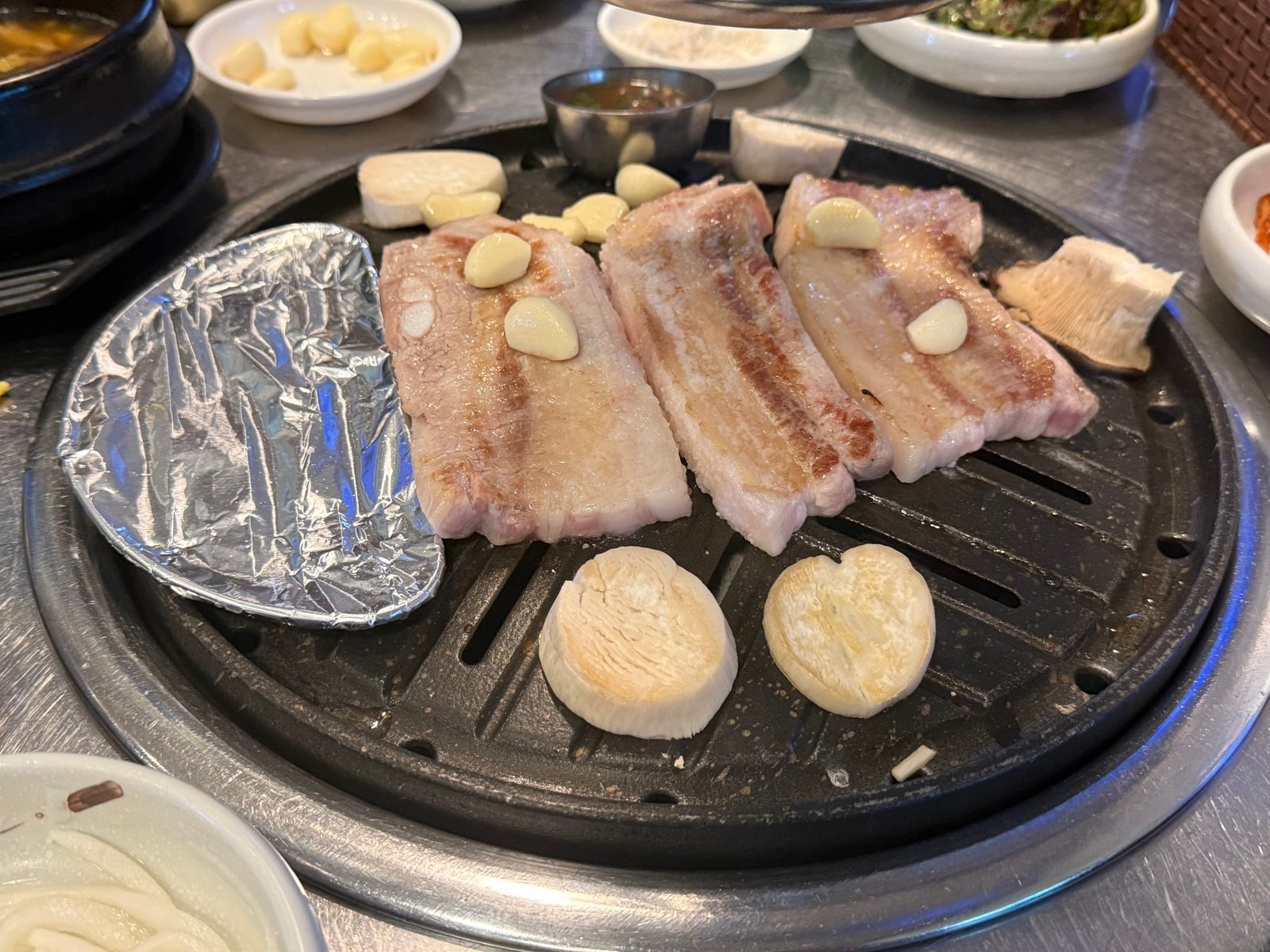 둥근 불판 위에 올린 두툼한 삼겹살 세 덩이와 마늘, 은박지