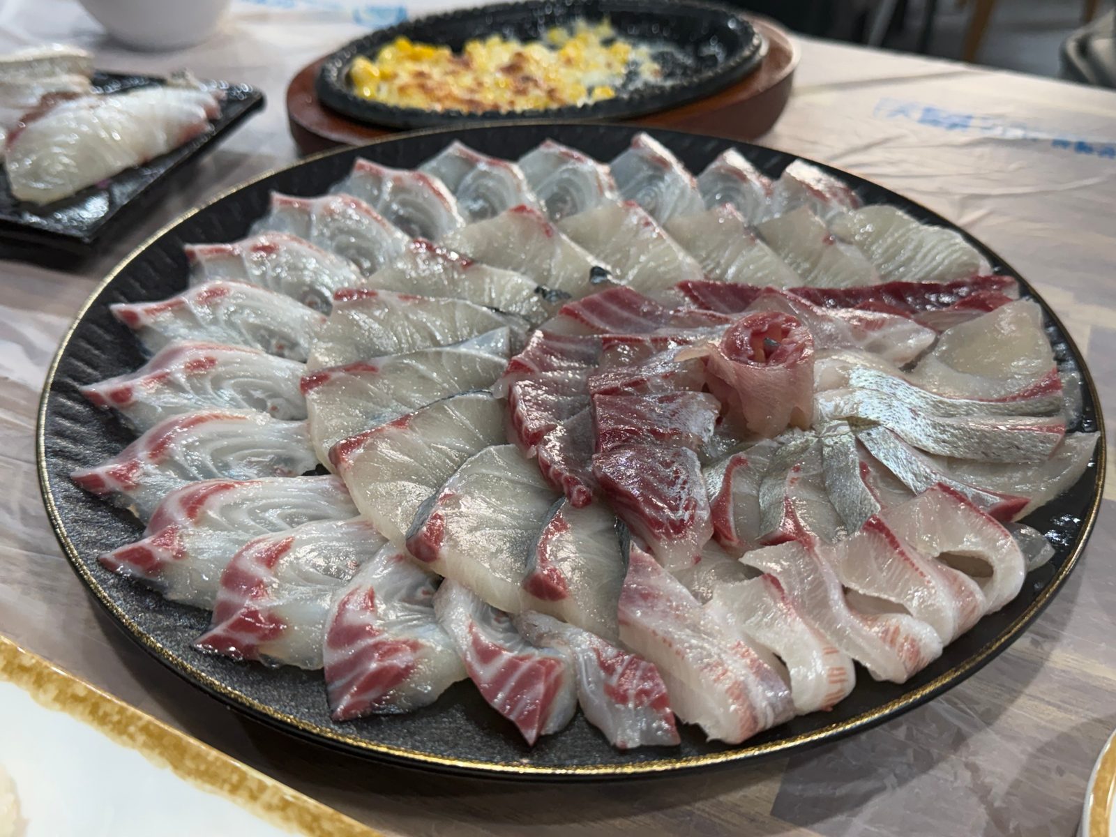 검은 접시에 꽃잎처럼 둥글게 담은 회와 뒤쪽의 콘치즈