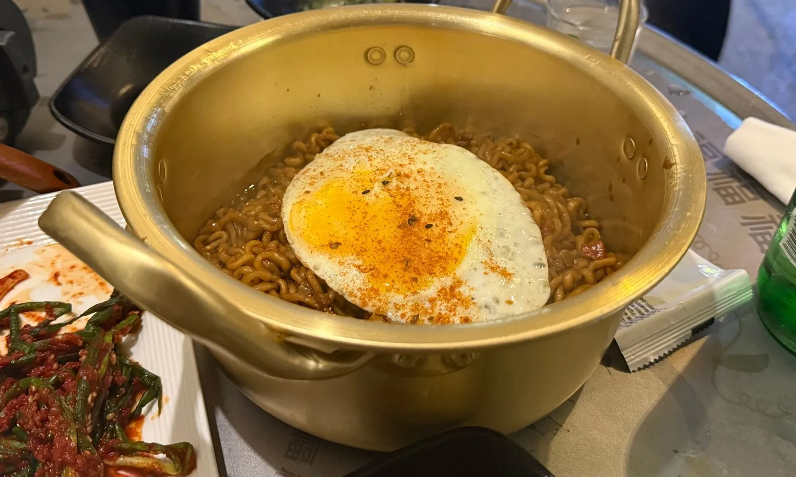 양은 냄비에 담긴 짜파게티와 그 위에 올린 달걀프라이, 옆에 놓인 파김치